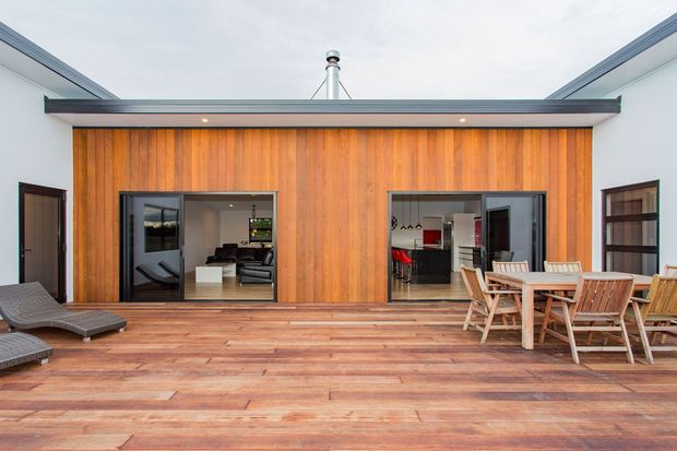 A contemporary courtyard-style outdoor deck enclosed by white walls, with a warm vertical timber-clad feature wall and two large glazed openings into the interior. The deck has extensive timber flooring, outdoor lounge chairs, and a timber dining setting. Interior living and kitchen areas are visible through the glazing, while a slim roof form and exposed flue add architectural definition.