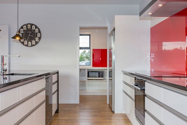 Contemporary galley-style kitchen with glossy white cabinetry, bold red glass splashback panels, dark stone benchtops, integrated stainless-steel appliances, timber flooring, and a pantry or utility area visible beyond the doorway. A large black wall clock and pendant lighting add contrasting details.