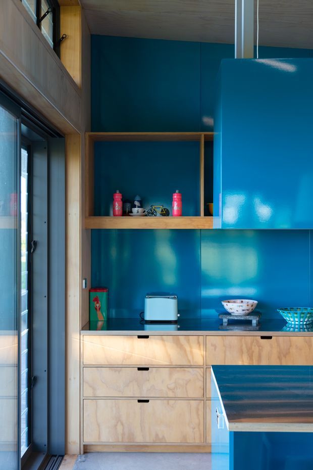 A compact, highly crafted kitchen or kitchenette with vivid glossy blue cabinetry and matching blue splashback walls, contrasted with light timber plywood drawer fronts and open timber shelving. A dark benchtop includes a toaster and decorative bowls, while a glazed external door and high-level windows bring in natural light. The composition highlights built-in joinery, strong colour blocking, and warm timber detailing.