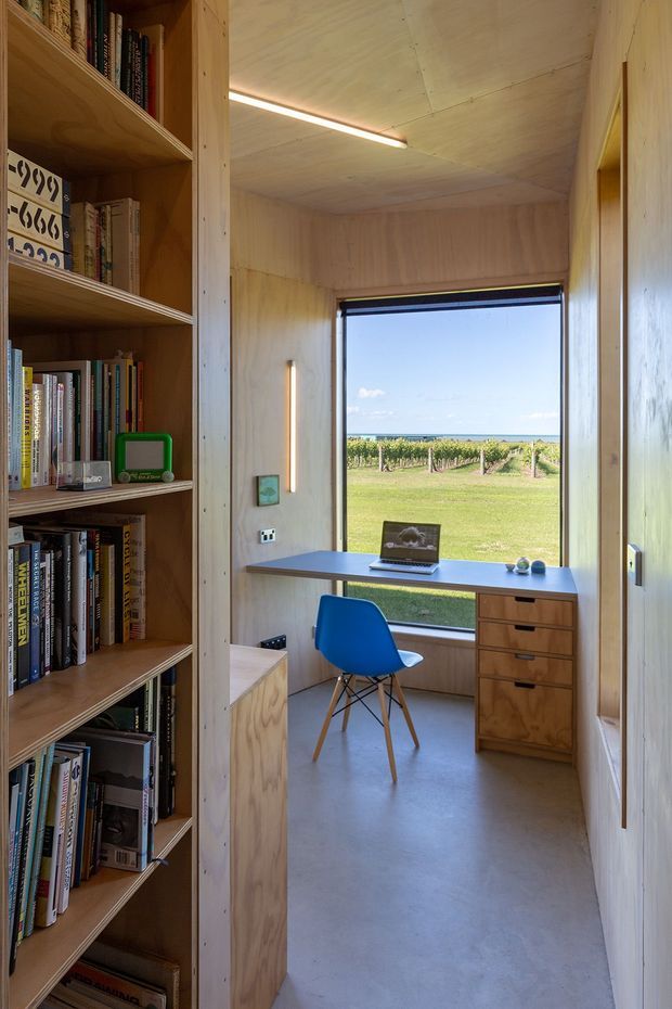 A compact timber-lined study nook viewed through a doorway, with built-in open bookshelves on the left, a wall-mounted desk and drawer unit beneath a large window, and a blue chair. The room has plywood or light timber wall and ceiling surfaces, a smooth grey floor, integrated linear lighting, and a rural vineyard landscape visible through the window.