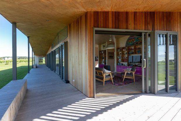 A covered exterior deck or verandah runs alongside a modern timber-clad building, with slim dark structural posts, extensive floor-to-ceiling glazing, and a large open sliding glass doorway. Through the opening is a colourful living room with timber shelving, a television, lounge chairs, and purple furnishings. The deck and soffit feature warm-toned timber, while strong sunlight creates striped shadows across the pale decking.
