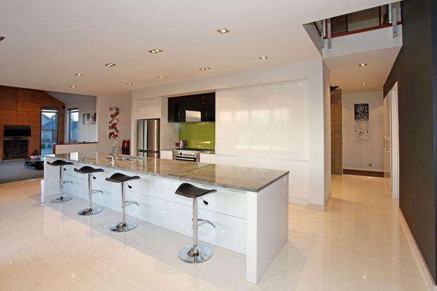 Bright open-plan contemporary kitchen with a large white island, polished stone benchtop, integrated cabinetry, green glass splashback, stainless-steel appliances, and five black bar stools. The kitchen opens toward a living area with a brick feature wall, while a hallway and upper-level void are visible to the right.