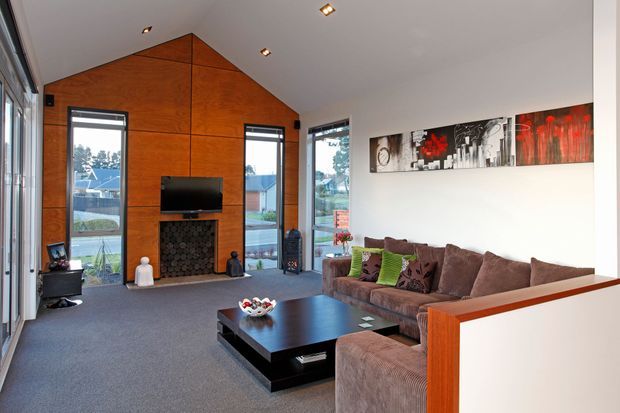Contemporary living room with a high pitched ceiling and full-height glazed doors and windows. A warm timber-panelled gable feature wall frames the fireplace and television, while a large sectional sofa, dark rectangular coffee table, recessed spotlights, and abstract wall art furnish the space. The room opens visually to an outdoor residential setting.