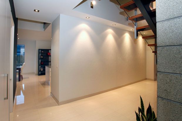A bright, minimalist interior hallway or open entry area with glossy light flooring, white textured walls, recessed spotlights, and a contemporary open-riser staircase with dark steel supports and timber treads. A partial concrete block feature wall is visible at right, with a glimpse of shelving and adjoining rooms beyond.