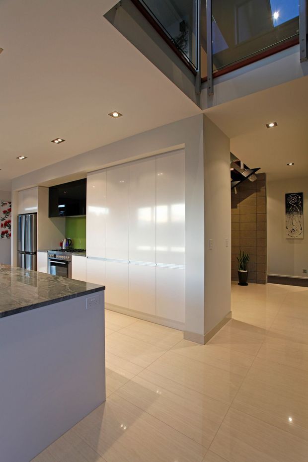 A bright contemporary open-plan kitchen viewed from the island, with glossy white full-height cabinetry, stainless-steel refrigerator and oven, a dark stone island benchtop, pale large-format tiled flooring, and recessed ceiling lighting. A stairway and double-height glazed void are visible beyond the kitchen.