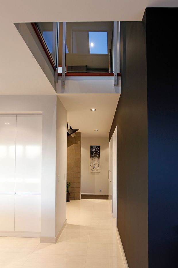 A clean contemporary interior hallway with pale tiled flooring, white walls and built-in white cabinetry. The corridor leads toward a tiled feature wall and artwork, while a glazed upper-level void or landing with skylight-like overhead glazing brings natural light into the space. A dark charcoal feature wall contrasts with the otherwise light palette.