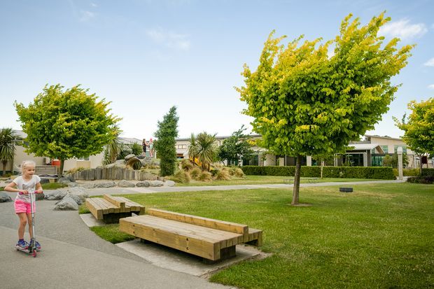A landscaped communal outdoor area with broad lawns, mature ornamental trees, native-style planting, low timber benches, and a paved winding pathway. A child on a scooter is present in the foreground, with low residential or community buildings visible beyond the landscaping.