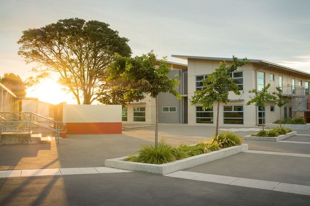 Contemporary multi-storey institutional or commercial building viewed from a landscaped paved forecourt at sunset. The building has a flat roof, light rendered walls, extensive horizontal glazing, balconies, and a clean geometric facade. Raised concrete planters with ornamental grasses and small trees define the foreground, while a low wall with a warm orange accent panel and accessible ramp/stair circulation are visible to the left.