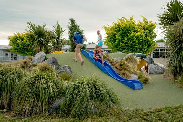 A landscaped outdoor play area with a small artificial-turf mound, bright blue slide, children playing, large decorative rocks, ornamental grasses, yucca-like plants and low shrubs. Low modern buildings are visible in the background.