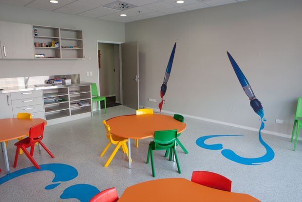 Bright children's activity or classroom space with small colorful tables and chairs, extensive built-in storage and shelving, a suspended acoustic tile ceiling, and playful blue floor and wall graphics. The room has a clean, functional institutional fit-out with vivid primary-color accents.