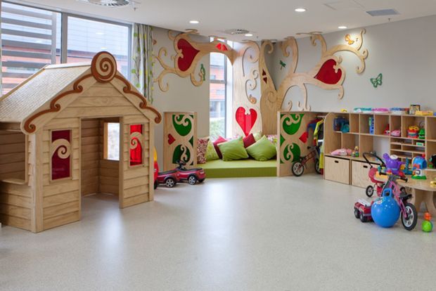 Bright, playful children's playroom with a whimsical fairytale mural, a wooden playhouse, colorful built-in shelving filled with toys, a window seat with cushions, and children's ride-on toys. The space has a light neutral floor, white ceiling, and abundant natural light.