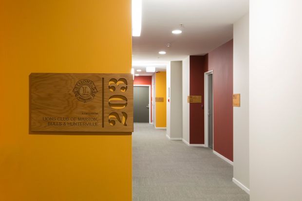 A clean institutional corridor with bright orange, red, and white walls, grey carpet, recessed ceiling lights, and multiple office or room doors. A large carved timber sign for the Lions Club of Marton and Bulls Health Centre is mounted prominently on the orange wall in the foreground.