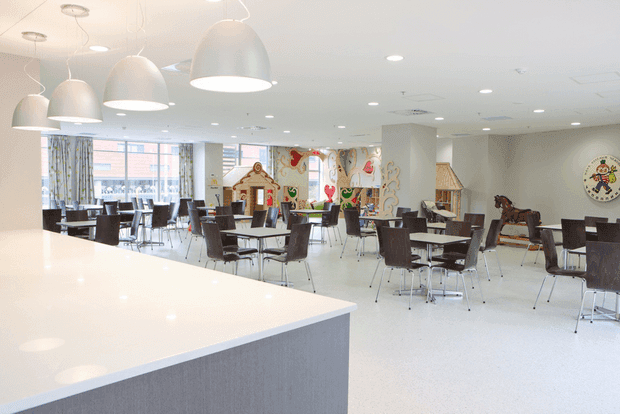 A bright, spacious commercial dining and activity area, likely in a childcare or family-oriented facility. The room features numerous dark tables and chairs, a large pale reception or service counter in the foreground, playful colourful wall murals and artwork, child-oriented furniture and toys, patterned curtains, and large pendant lights beneath a white acoustic ceiling.