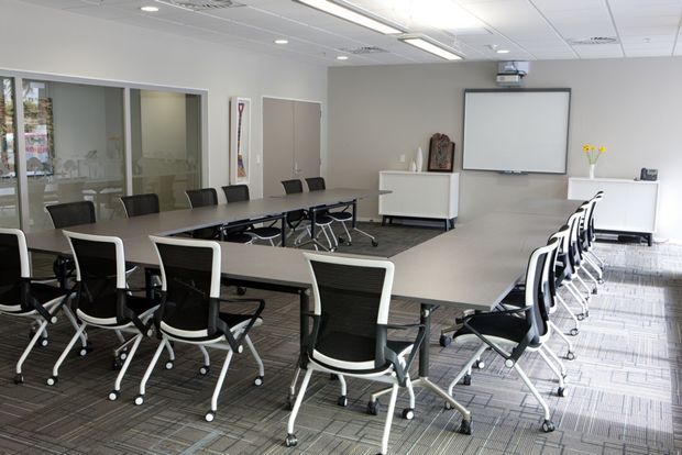 A bright contemporary commercial conference or training room with large modular tables arranged in a U-shape, numerous black-and-white wheeled office chairs, a wall-mounted projection screen, recessed ceiling lighting, carpet tile flooring, and glazed partitions along one side.