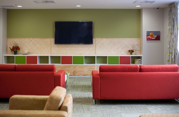 A colourful contemporary lounge or media room viewed from behind red sofas. The room features a wall-mounted television, a green feature wall, pale patterned wall panelling, and a long built-in display and storage unit with alternating red, green and white compartments. Neutral patterned carpet, recessed ceiling lights, artwork and curtains complete the space.
