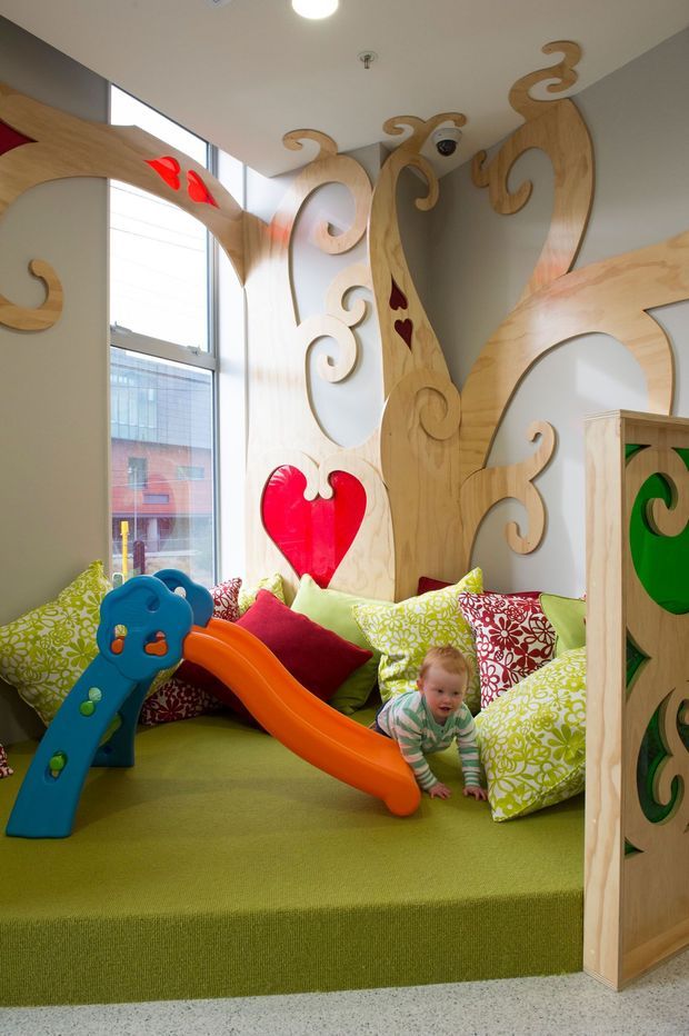 A bright, playful children's playroom with a raised green carpeted platform, colourful patterned cushions, and a small plastic slide. The room features elaborate custom timber wall and ceiling panels with curling organic cut-out motifs, including red and green decorative inserts. Large windows provide abundant natural light, and a crawling child is visible near the slide.