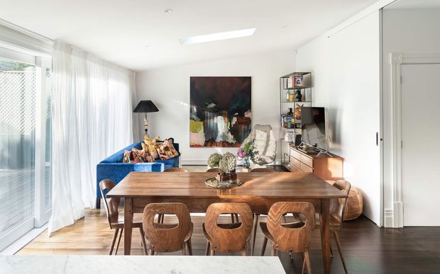 Bright open-plan dining and living area with a substantial timber dining table and sculptural wooden chairs in the foreground. The room has white walls and ceiling, light timber flooring, sheer floor-to-ceiling curtains, large glazed doors, a blue sofa, artwork, shelving, and warm contemporary furnishings.