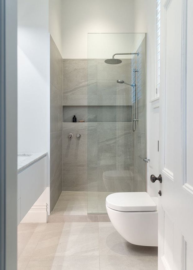Bright, narrow contemporary bathroom with a walk-in tiled shower, frameless glass screen, recessed shower niche, wall-mounted rainfall and handheld fixtures, and a wall-hung white toilet. Large pale grey stone-look tiles cover the shower walls and floor, while a white paneled door and plantation-shuttered window add traditional detailing.