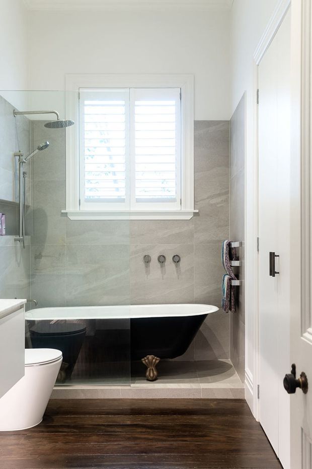 Bright, compact traditional bathroom featuring a freestanding black clawfoot bathtub, glass-enclosed shower, wall-mounted shower fixtures, a white toilet, pale stone-look wall tiles, white plantation shutters, and dark timber flooring. White painted trim and cabinetry/door hardware add contrast.