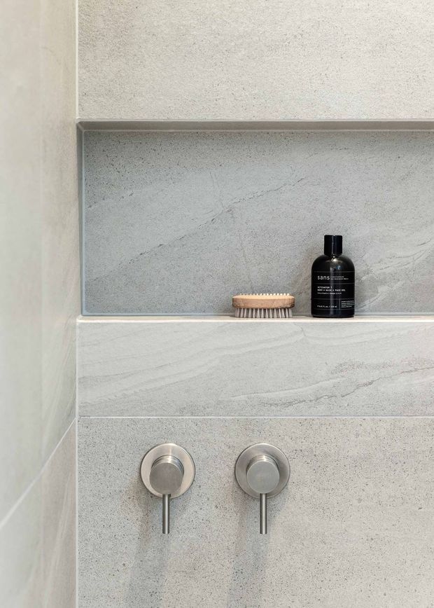 Close-up of a minimalist bathroom shower wall featuring pale grey textured stone or concrete-look surfaces, a recessed niche with a small brush and black bottle, and two round brushed-metal shower controls.