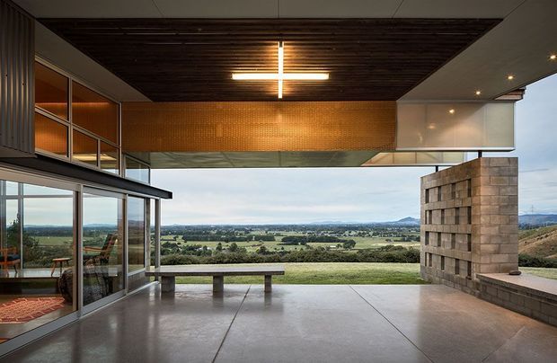 A dramatic covered outdoor terrace or viewing pavilion opens toward a broad rural landscape. The space features a polished concrete floor, a dark timber-lined recessed ceiling with a modern cross-shaped light, expansive glazed walls, a warm copper-toned wall panel, exposed concrete block construction, and built-in concrete bench seating. The architecture has a strong indoor-outdoor connection and a cantilevered, open-plan composition.