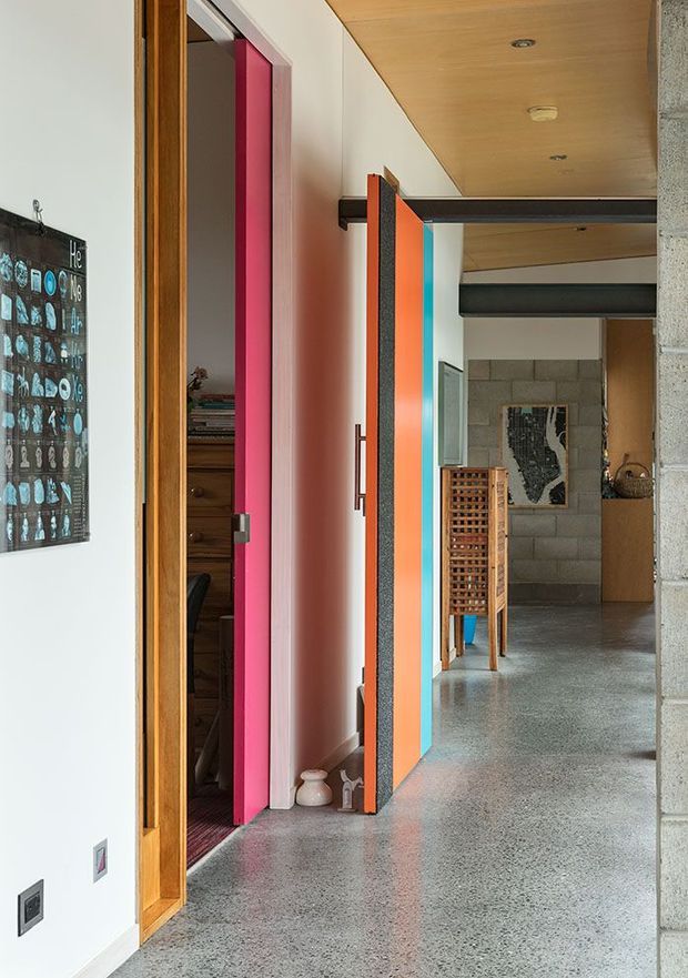Interior view down a colourful residential hallway, with polished grey terrazzo flooring, white walls, warm timber ceiling panels, exposed dark structural beams, and vivid turquoise, orange, and magenta doors. A home office or study is visible through the open doorway on the left, while the hall leads toward a kitchen or living area with exposed concrete block walls and timber joinery.