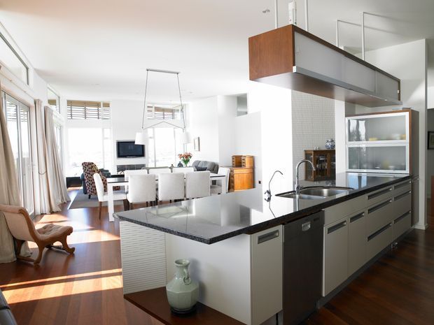 Bright open-plan kitchen and dining/living area with a large dark stone island, white fixed cabinetry, integrated appliances, and a suspended timber-and-glass overhead cabinet. The space features polished timber flooring, white walls, extensive glazing, and contemporary mid-century-inspired furnishings.