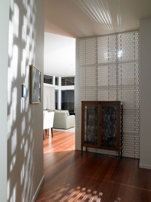 Interior view of a bright contemporary residential hallway or entry area opening into a living room. A full-height perforated breeze-block screen creates a patterned privacy divider behind a dark timber glass-front cabinet. Rich dark timber flooring, white walls, high glazing and strong filtered sunlight define the space.