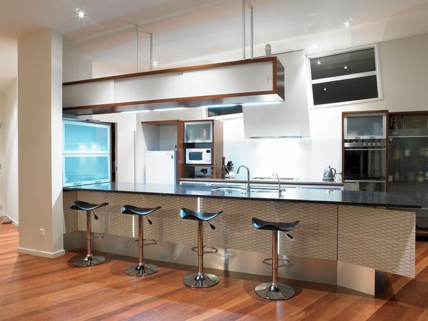 Modern open-plan kitchen dominated by a large island with a dark stone benchtop, integrated sink and five contemporary bar stools. The island has textured horizontal cabinetry, while the surrounding kitchen features white cabinetry, stainless-steel appliances, timber flooring, pendant-style overhead joinery, and recessed lighting. A dining or living area is partially visible beyond the kitchen.