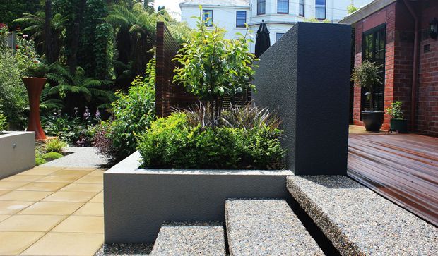 A landscaped contemporary courtyard or terrace with rendered geometric planter walls, gravel-covered stepped seating or stairs, timber decking, lush planting, and a paved outdoor area. A red-brick building and white multi-storey building are visible in the background.