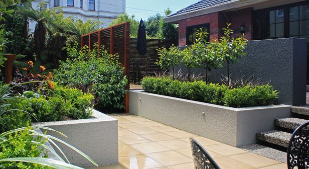 A landscaped courtyard or patio beside a modern brick-clad building, featuring raised rendered planter beds, dense layered planting, timber privacy screens, tan paved flooring, and outdoor seating. The space is bright and verdant with a private urban-garden atmosphere.