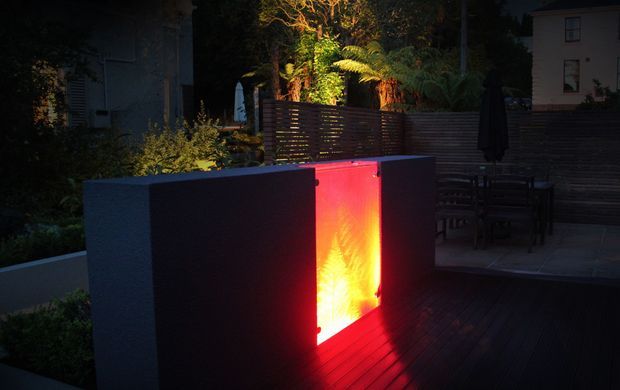 Moody evening view of a landscaped outdoor terrace with dark timber decking, horizontal timber screening, outdoor dining furniture, and a dramatic illuminated red-orange feature panel set within a low rendered wall. Dense planting and a fern are visible behind the screen.