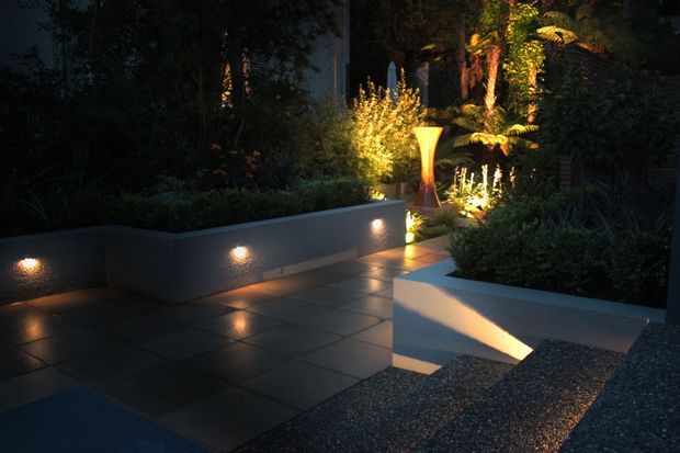 A moody landscaped courtyard or garden at night, featuring dark tiled paving, raised rendered planter beds, dense planting, integrated wall and path lighting, and illuminated steps leading through the garden. A sculptural warm-toned outdoor light or feature stands among the vegetation.