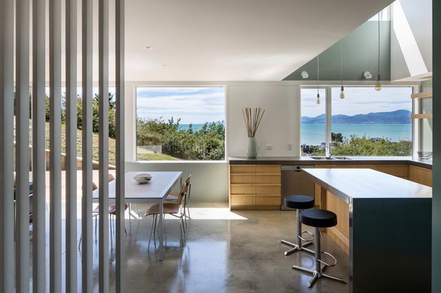 A bright contemporary open-plan kitchen and dining area with expansive views toward blue water and hills. The kitchen features warm timber cabinetry, a large dark island with a pale benchtop, pendant lights, polished concrete flooring, and a dramatic angled glazed ceiling or clerestory element. Vertical timber screening partially separates the dining space, while large windows bring in abundant natural light.