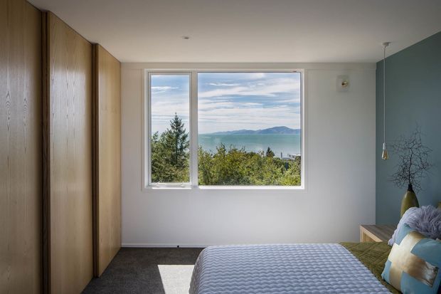 A calm, compact bedroom with a large picture window overlooking water and distant hills. The room features full-height natural timber wardrobe cabinetry along the left wall, a grey carpeted floor, white walls and ceiling, and a muted blue-grey feature wall. A neatly made bed with a textured grey cover and layered blue, green and cream cushions occupies the foreground, with a small timber bedside table and pendant light at right.