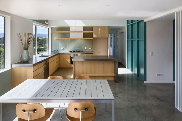 A bright contemporary open-plan kitchen and dining area. The kitchen features light timber cabinetry, open timber shelving, a dark stone island benchtop, integrated appliances, and a teal built-in shelving or partition wall. A white dining table with timber chairs occupies the foreground. Polished concrete flooring, large windows, and a mirrored splashback create a clean, airy space with views to the landscape outside.
