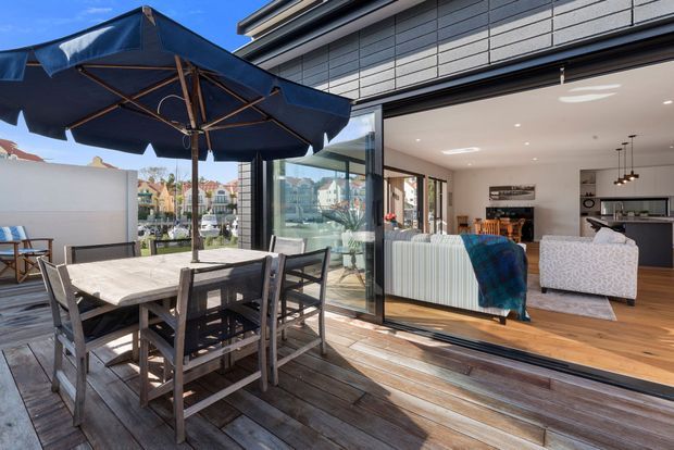 A bright contemporary home viewed from a timber outdoor deck, with a large navy patio umbrella shading an outdoor dining table and chairs. Fully open sliding glass doors connect the deck to a warm timber-floored living room with a sofa, armchairs, pendant lights and a compact kitchen. The exterior facade features dark horizontal cladding and extensive glazing, with a waterfront-style residential setting visible beyond.