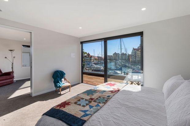 Bright bedroom with a white bed, textured white bedding, and a colourful patchwork runner. The room has pale neutral walls, carpeted flooring, recessed ceiling lights, and large black-framed sliding glazing opening to a marina view. A small chair sits near the window, with a glimpse of an adjacent furnished room through the doorway.