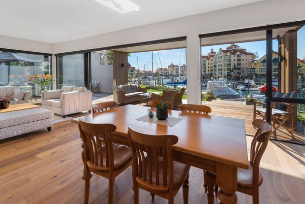 Bright open-plan dining and living area with a large timber dining table and carved wooden chairs in the foreground, upholstered lounge seating to the side, and extensive dark-framed glazing overlooking a marina and waterfront apartments. Warm timber flooring and furnishings create an inviting, airy interior.