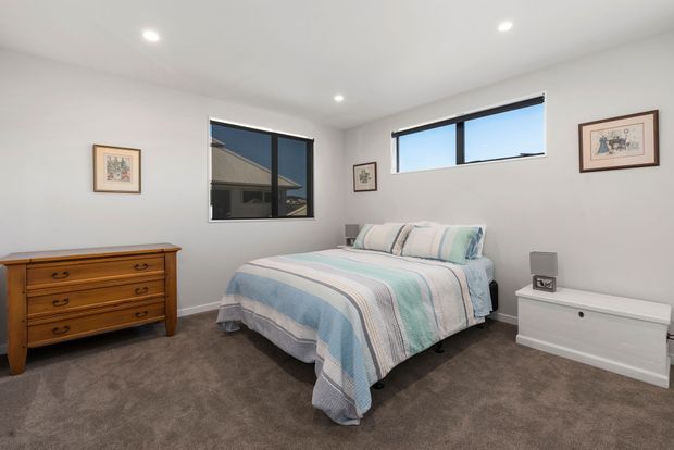 Bright, simply furnished bedroom with a double bed dressed in a soft striped blue, grey and white bedspread, a timber chest of drawers, a white low storage unit, framed artwork, recessed ceiling lights, carpeted flooring, and large dark-framed windows.