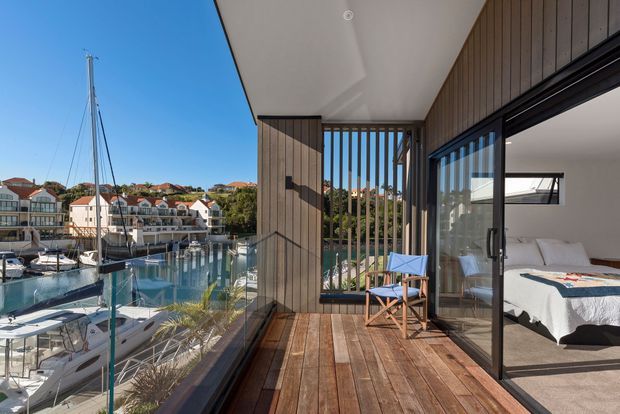 A bright waterfront balcony or terrace overlooking a marina, with timber decking, vertical timber screening, full-height glass balustrades and dark-framed sliding doors. A bedroom with a white bed is visible through the glazing, while boats and neighbouring homes sit beyond the water.