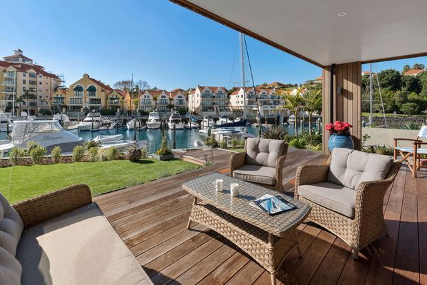 A bright waterfront outdoor living area on a timber deck overlooking a marina. Wicker lounge seating, a glass-topped coffee table, potted plants, and a manicured lawn sit beneath a broad covered roof, with boats and apartment-style buildings visible across the water.