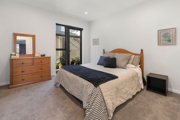 A bright, contemporary bedroom with a wooden bed featuring turned posts and a curved headboard, coordinated patterned bedding in neutral tones with navy accents, a timber dresser and mirror, grey carpet, white walls, recessed ceiling lighting, and a full-height glazed door opening to an outdoor area.