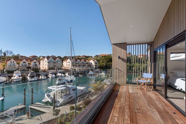 A sunny waterfront apartment balcony overlooking a marina, with a timber deck, clear glass balustrade, vertical timber screening and wide sliding glass doors opening into a bedroom. The room beyond contains a bed, while boats and waterfront buildings are visible outside.