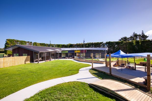 A low-rise contemporary community or educational building set within landscaped grounds. The dark pitched roof and timber-clad wing open onto a broad lawn, with glazed façades, a curved concrete footpath, timber boardwalk, fenced play area, and shade canopies visible in the foreground.