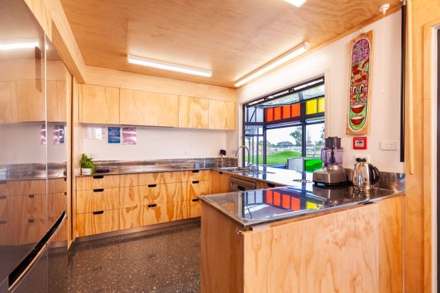 Compact contemporary kitchen with extensive light plywood cabinetry, matching plywood ceiling and wall panelling, dark polished benchtops, and a central island. A colorful glazed window provides a strong accent and overlooks a green outdoor setting. The space includes integrated appliances, open shelving, a sink, cooktop, and small appliances.