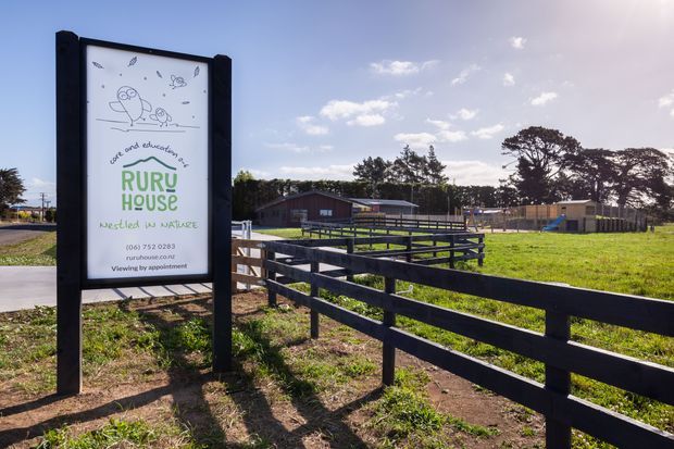 A sunny outdoor view of a rural childcare property entrance, with a large illustrated sign for Ruru House nestled in nature, dark timber fencing and accessible ramped walkways leading toward low timber buildings and a playground. The scene has open grassy grounds, mature trees, and a clear blue sky.