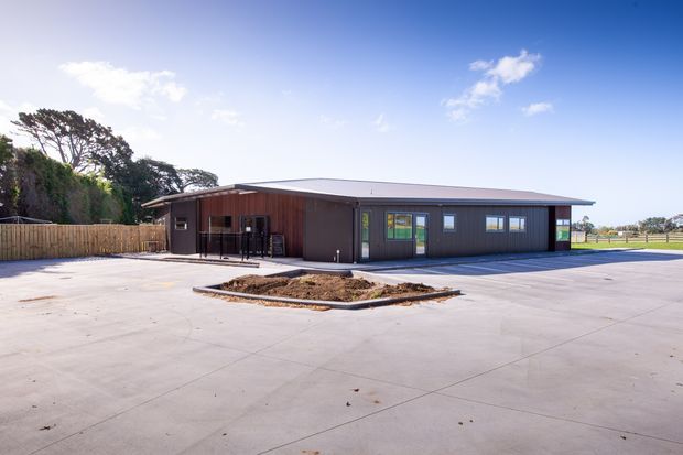 Single-storey modern low-rise building with dark vertical cladding, a shallow pitched or low-slope metal roof, large paved forecourt, and a small landscaped planter. The building appears institutional or commercial, with multiple glazed doors and windows, outdoor seating near the entrance, fencing, and open grounds surrounding it.