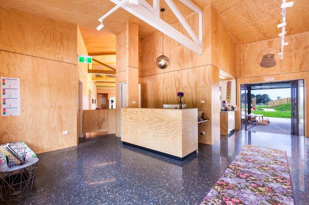 A bright contemporary reception or entry lobby lined with warm plywood walls and ceilings. A built-in plywood reception desk sits centrally on a polished dark terrazzo floor, with white track lighting and exposed structural elements overhead. A glazed doorway opens to a landscaped outdoor area, while a colourful floral rug and small seating area add visual warmth.