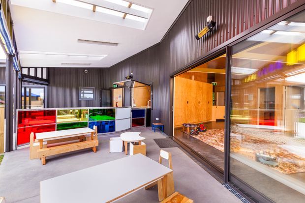 A bright, contemporary communal interior with a concrete floor, dark vertically ribbed wall cladding, skylights, and extensive internal glazing. Low timber tables and benches sit beside colourful built-in storage or display units, while a large glazed opening reveals a warm timber-lined adjoining activity room. The space has an institutional or educational character.